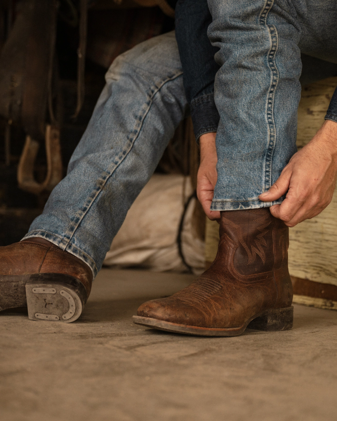 Person sitting, adjusting the cuff of blue jeans over worn brown cowboy boots in a rustic setting.