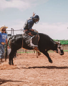A bull rider wearing protective gear rides a bucking bull in a rodeo arena while a person in a hat stands nearby watching.