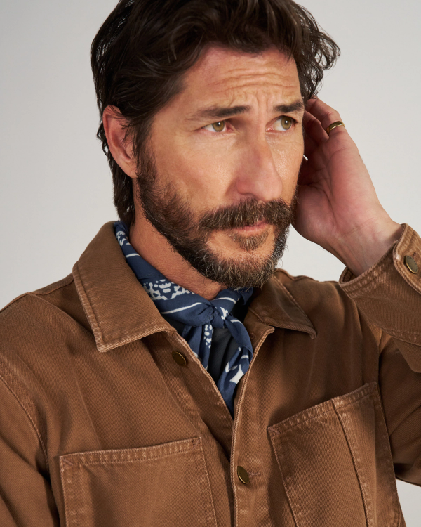 A man with dark hair and a beard wears a brown jacket and patterned blue scarf, looking concerned while touching the side of his head.