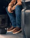 Person wearing brown boots, jeans, and a brown jacket sits on the edge of an open vehicle door.