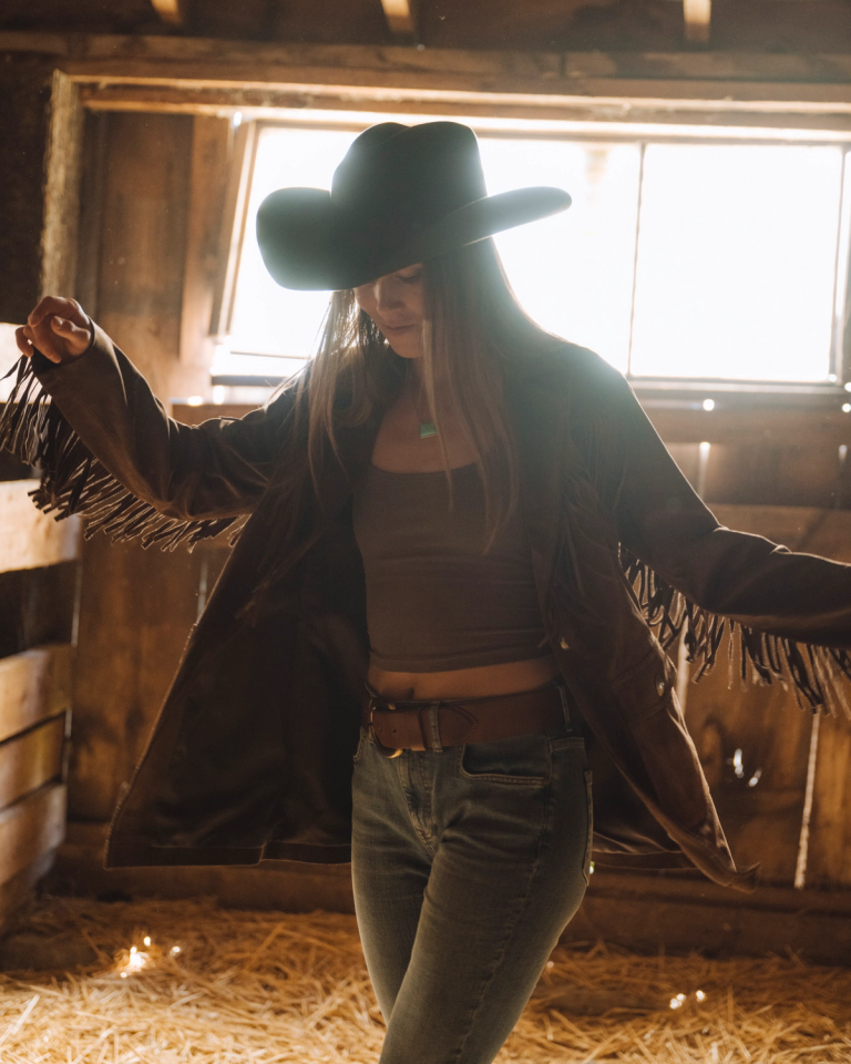 woman in dark suede fringe jacket, cowboy hat, and western belt dancing in a barn.