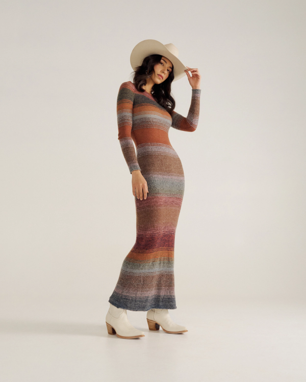 Woman wearing a striped, long-sleeve, fitted maxi dress, cream cowboy hat, and cream ankle boots standing against a plain background.
