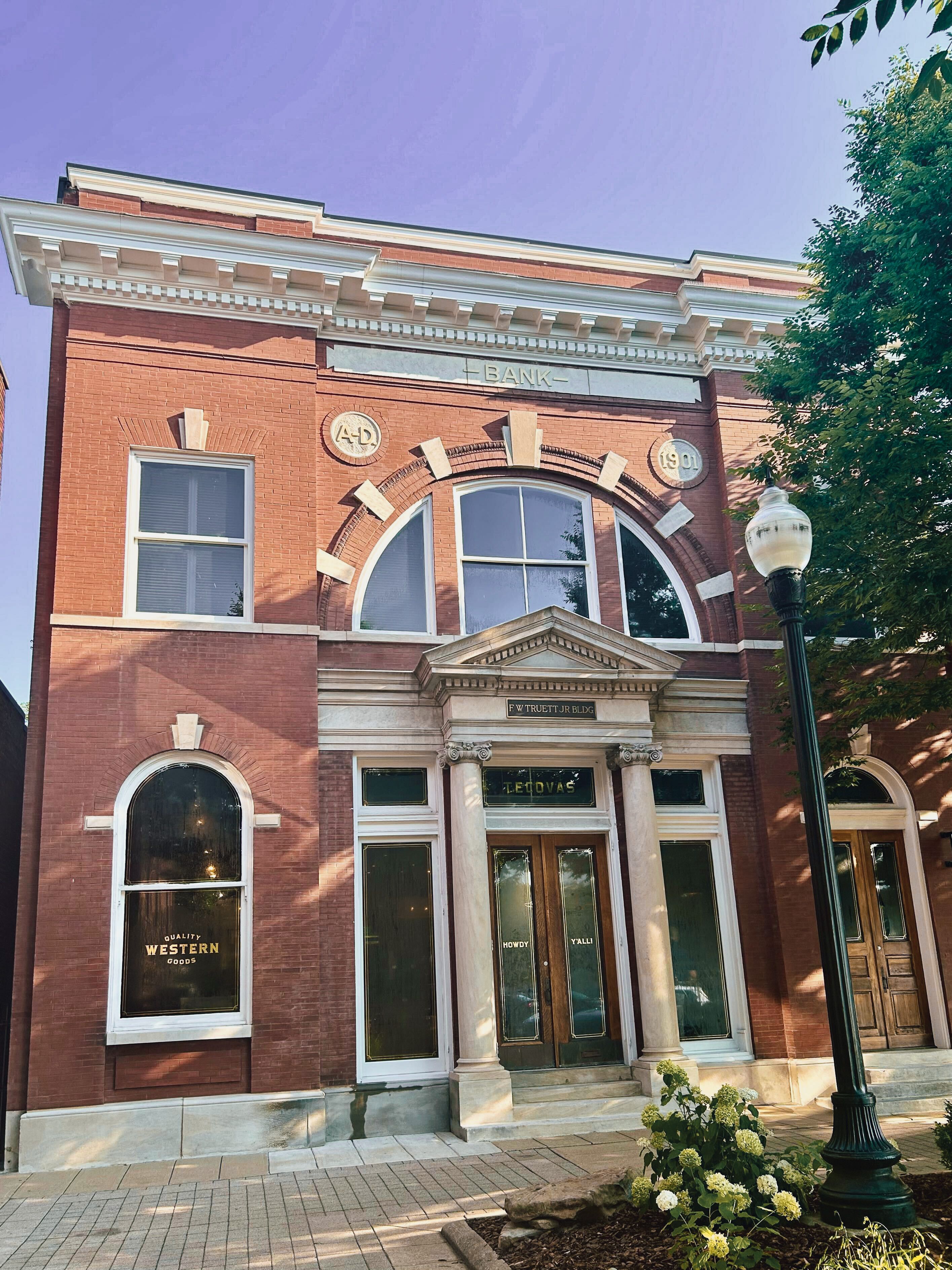 A historic red brick building with arched windows, a sign reading "BANK" above the entrance, and a streetlamp in front.