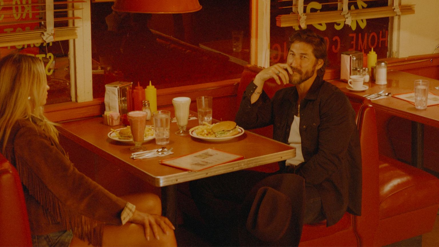Two people sit in a red booth at a diner with food and drinks on the table, under warm lighting. One person holds a hat on their lap.
