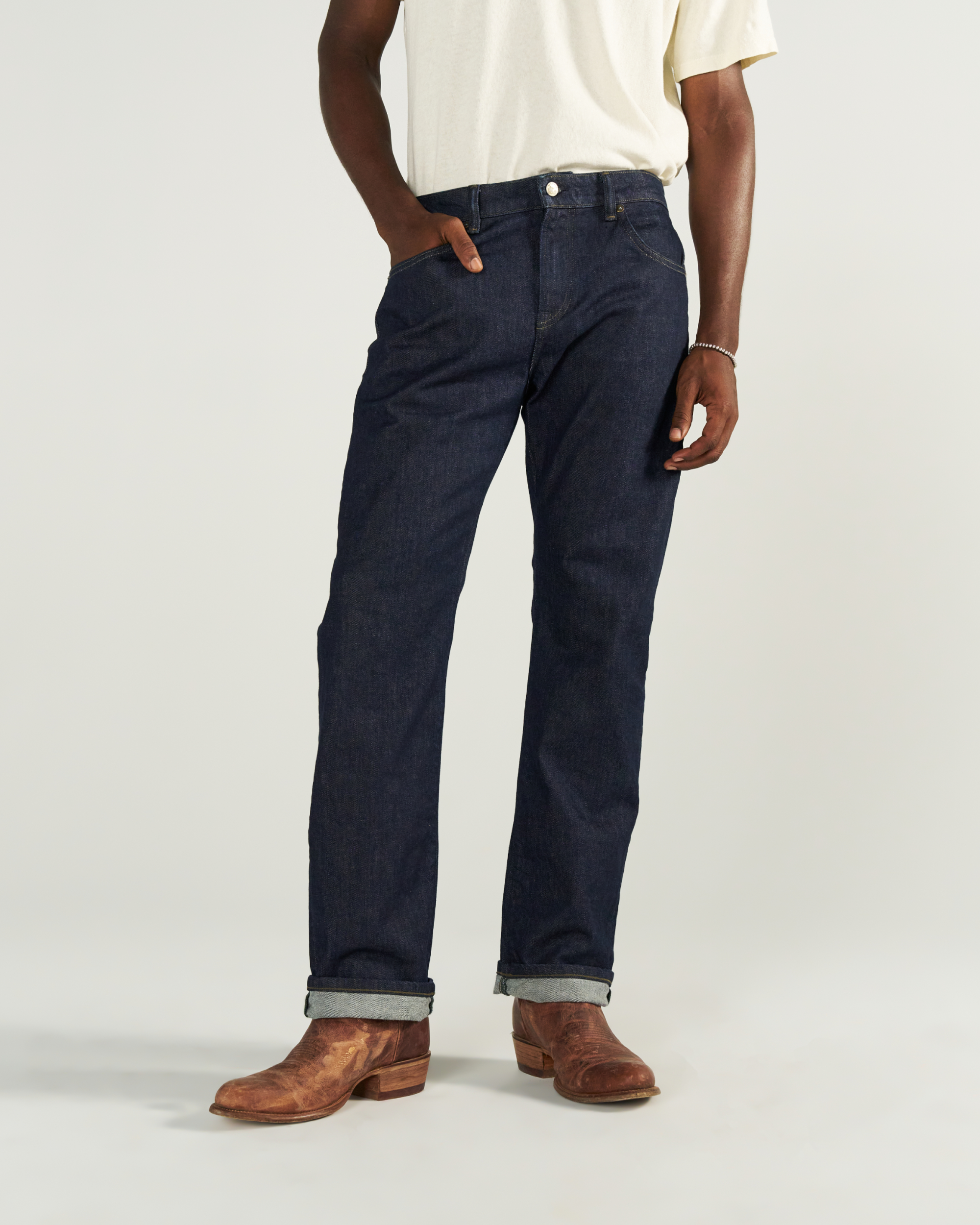 Person wearing a white t-shirt, dark blue jeans with rolled cuffs, and brown boots standing against a plain background.