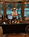 A person wearing a cowboy hat stands behind a table displaying various cowboy boots and accessories inside a rustic room with large windows.