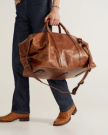 A person wearing dark jeans and tan boots holds a large brown leather duffel bag with handles and a shoulder strap.