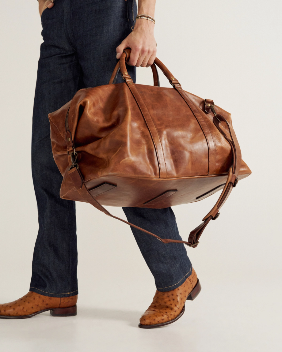 A person wearing dark jeans and tan boots holds a large brown leather duffel bag with handles and a shoulder strap.