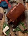 A brown leather toiletry bag on a wooden surface surrounded by decorative green ornaments, evergreen leaves, a blue hat with a red ribbon, and miniature cowboy boots.