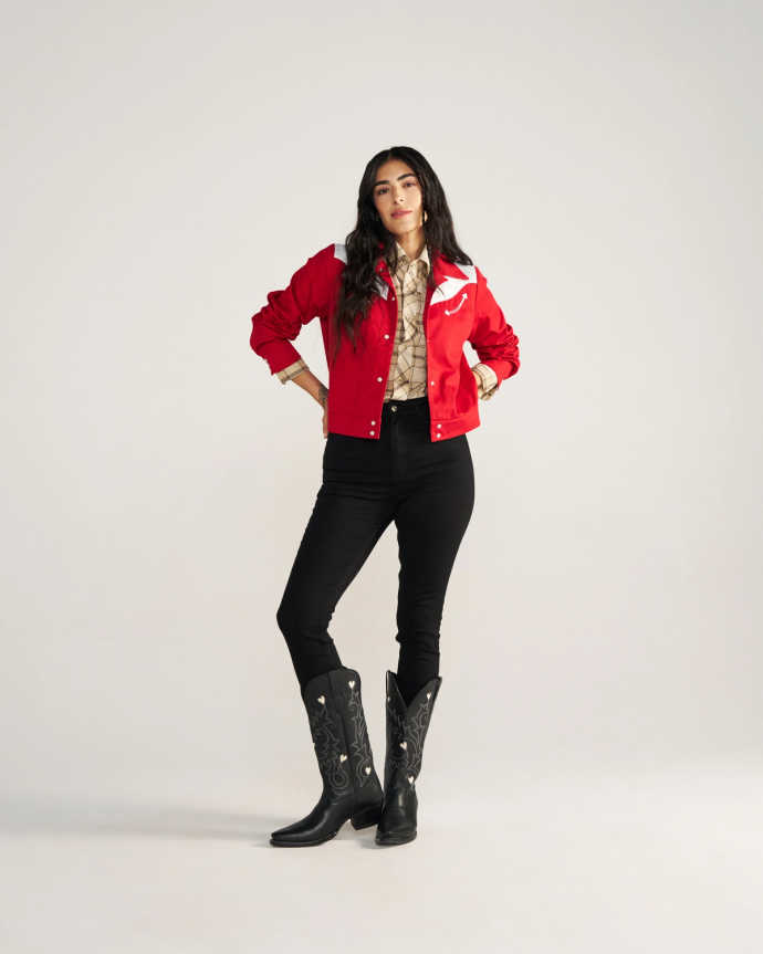 A person stands facing forward wearing a red jacket, patterned shirt, black pants, and black cowboy boots against a plain light background.