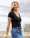 A person with long, blonde hair wearing a black short-sleeved shirt and blue jeans, standing outdoors against a background of a cloudy sky and bleachers.