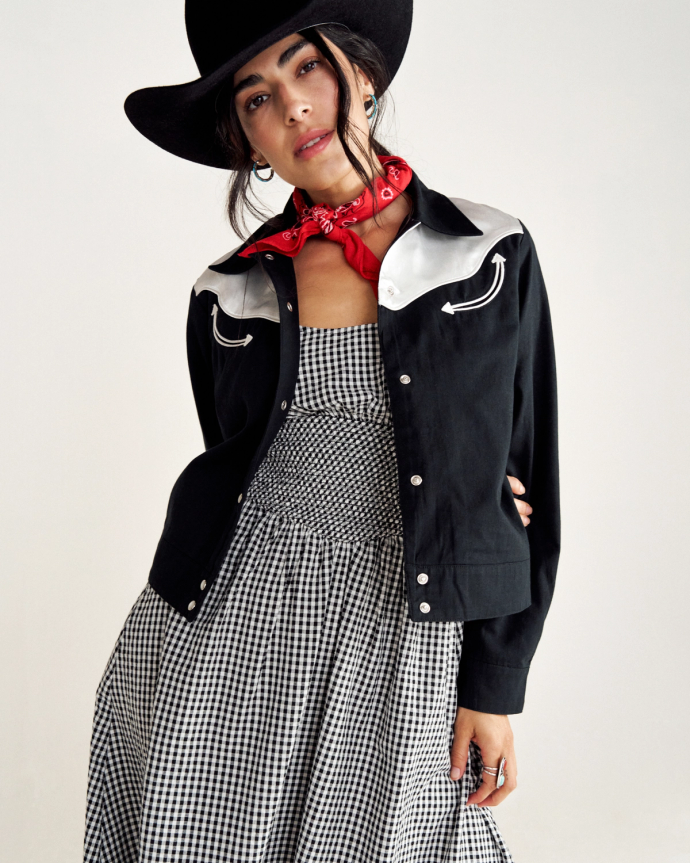 Person in a black cowboy hat and jacket, wearing a red bandana and a checkered dress, posed against a plain background.