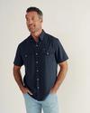 A man with short hair and a beard wears a navy short-sleeve button-up shirt and light blue jeans, standing against a plain off-white background.