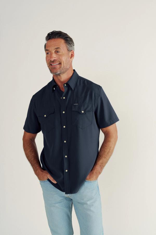 A man with short hair and a beard wears a navy short-sleeve button-up shirt and light blue jeans, standing against a plain off-white background.