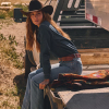 Woman in a cowboy hat and denim outfit sits on the back of a truck next to a wool blanket on a sunny day.