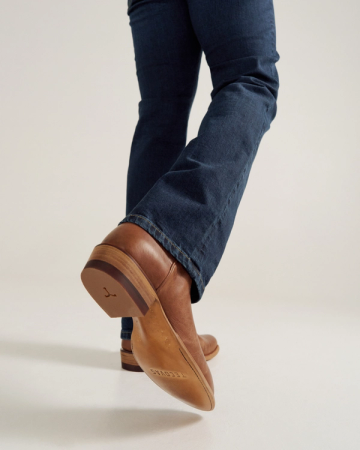 Person wearing blue jeans and brown leather shoes, photographed from behind at ankle level, with one foot slightly raised, showing the sole.