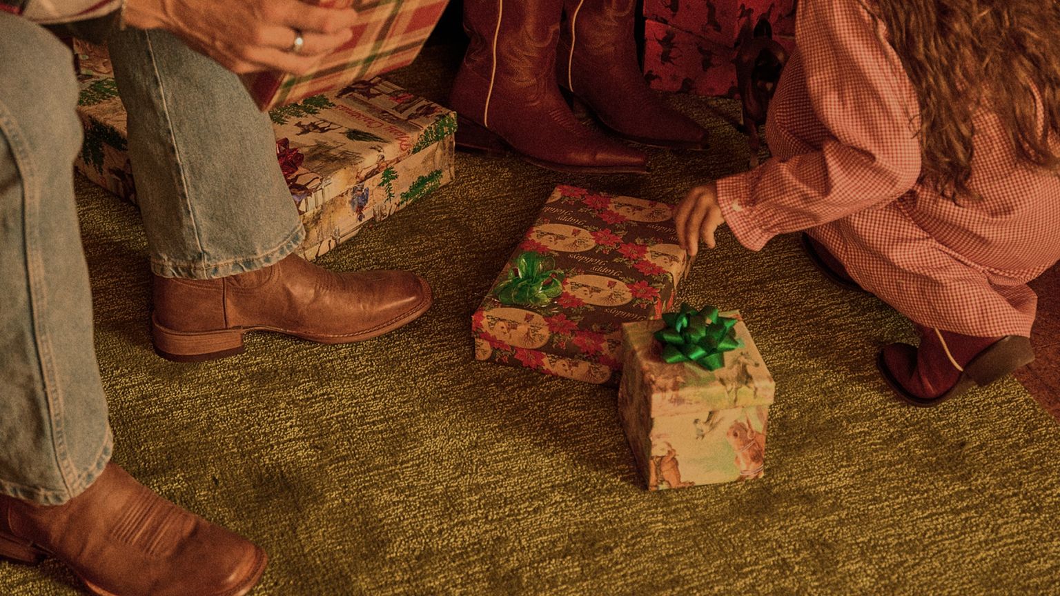 People wearing boots are sitting or kneeling on a green carpet, surrounded by wrapped gift boxes with bows.