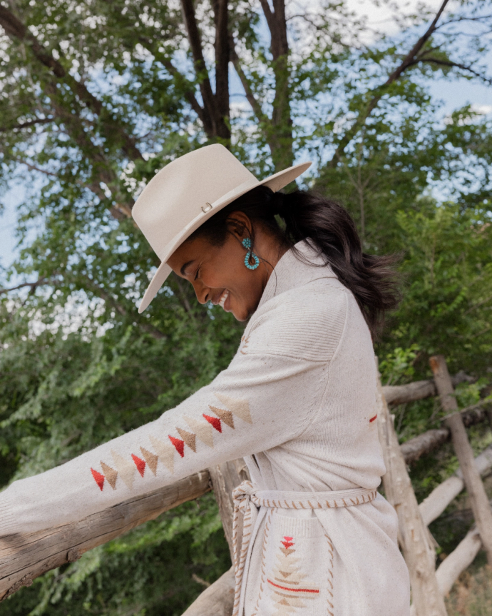 A woman wearing a light-colored hat and jacket with embroidered patterns leans on a wooden fence, set against a backdrop of green trees.