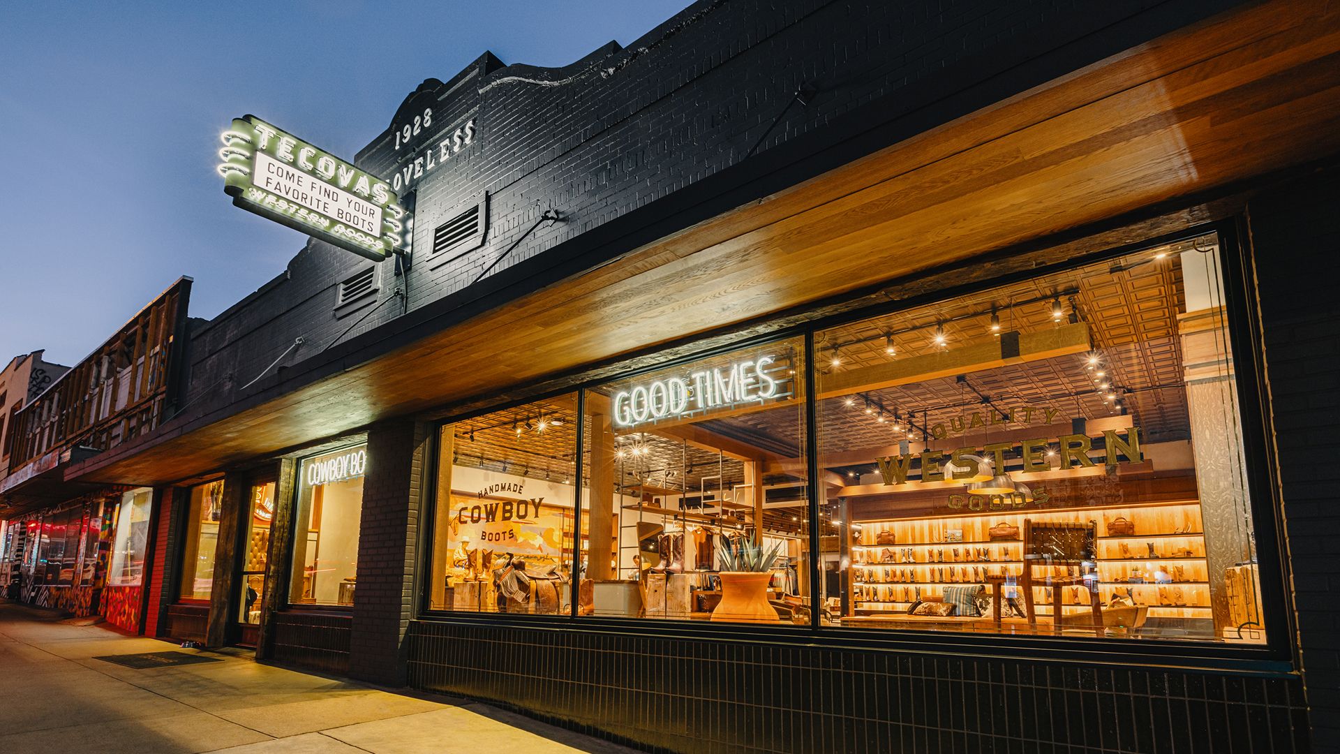 Storefront of Tecovas at dusk with illuminated signs reading "Good Times" and "Western Goods," showing boots and hats displayed inside through large windows.
