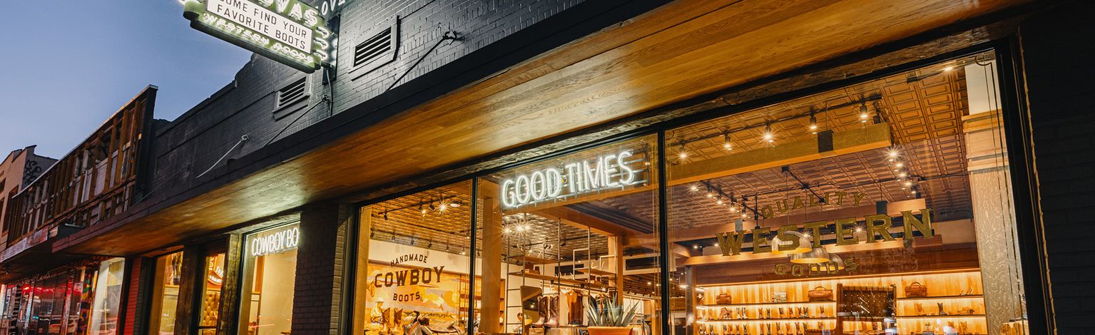 Storefront of Tecovas at dusk with illuminated signs reading "Good Times" and "Western Goods," showing boots and hats displayed inside through large windows.