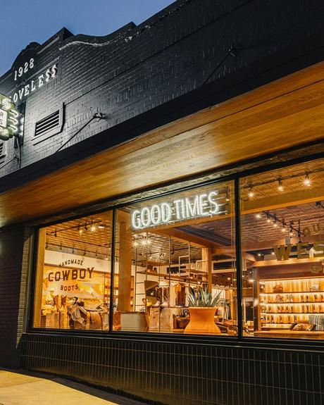 Storefront of Tecovas at dusk with illuminated signs reading "Good Times" and "Western Goods," showing boots and hats displayed inside through large windows.