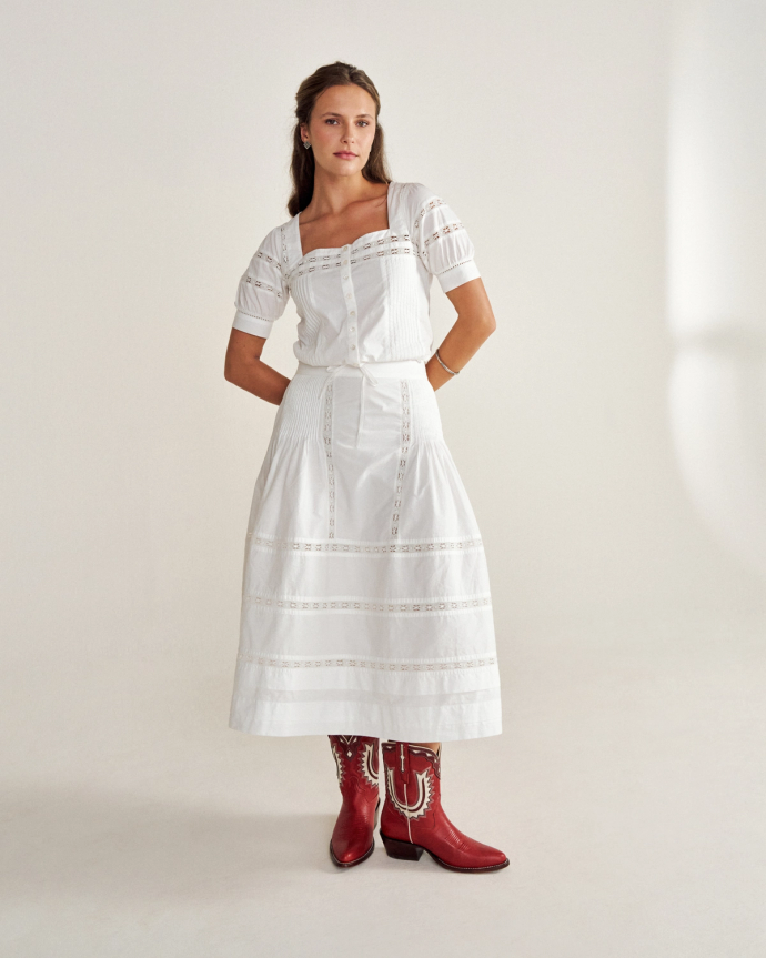 Person in white, lace-trimmed dress and red cowboy boots stands against a plain background.