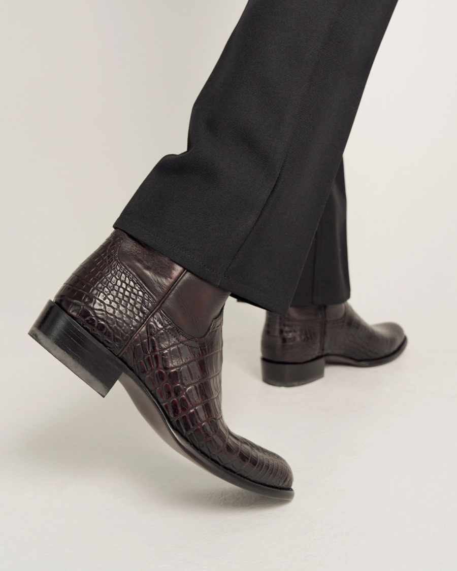 A person wearing black pants and dark brown crocodile-patterned leather ankle boots taking a step forward on a light-colored surface.