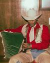 Person in cowboy attire, including a white hat and red shirt, sits casually on a brown sofa with a drink, next to a green pillow, against a wood-paneled wall.
