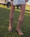 Person wearing The Sadie cowboy boots and a white lace dress stands on green grass with a vehicle in the background.