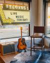 A stage area with an amplifier, an acoustic guitar, a tall chair, and a sign reading "Tecovas Live Music Tonight!" by large windows.