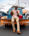 A person in casual clothes and a hat sits on the tailgate of a blue truck holding a football, with a cooler and orange decorations in the background.