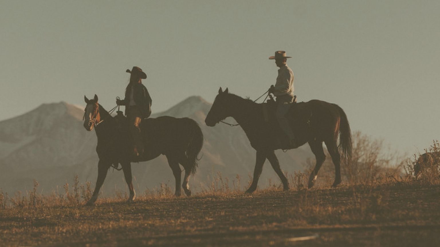 Two people wearing cowboy hats ride horses across an open field with mountains visible in the background.