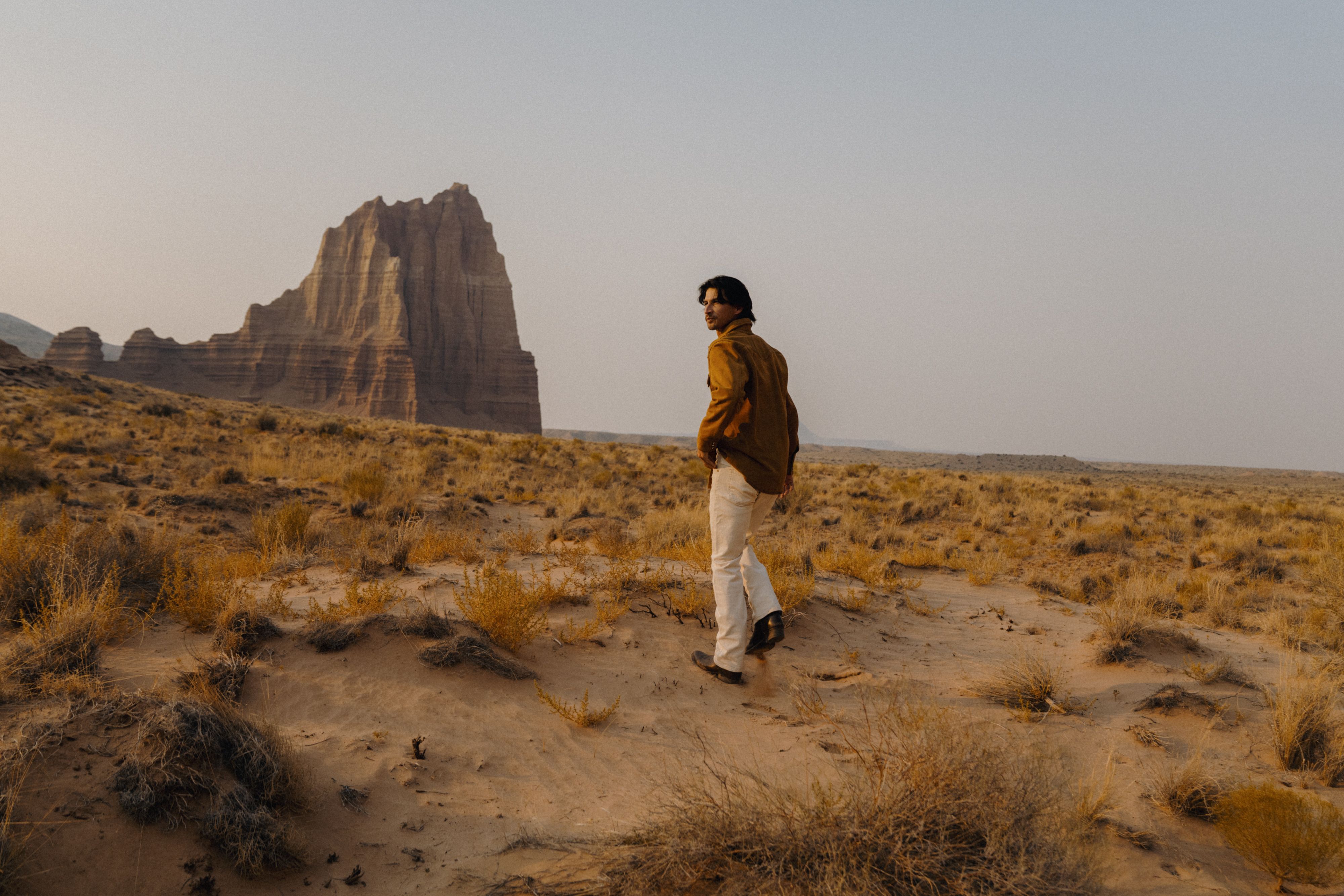 A person in a brown shirt and light pants walks through a desert landscape toward a large rock formation.