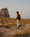 A person in a brown shirt and light pants walks through a desert landscape toward a large rock formation.