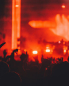 A crowd with raised hands is silhouetted against a red-lit stage with bright lights during a concert.