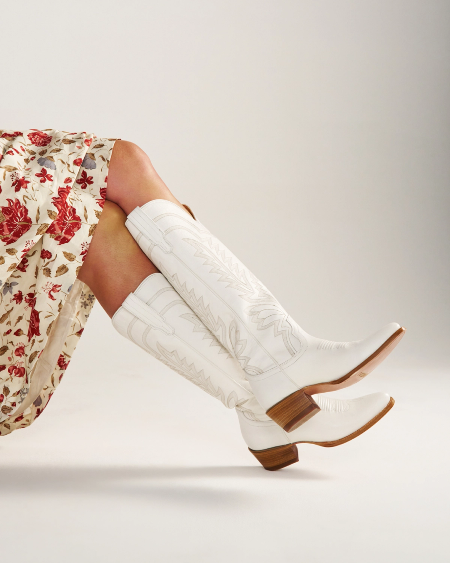 The Abby Wide Calf in Snow Cowhide. Person wearing a floral skirt and Snow cowhide white cowboy boots with brown heels; legs crossed against a plain light background.