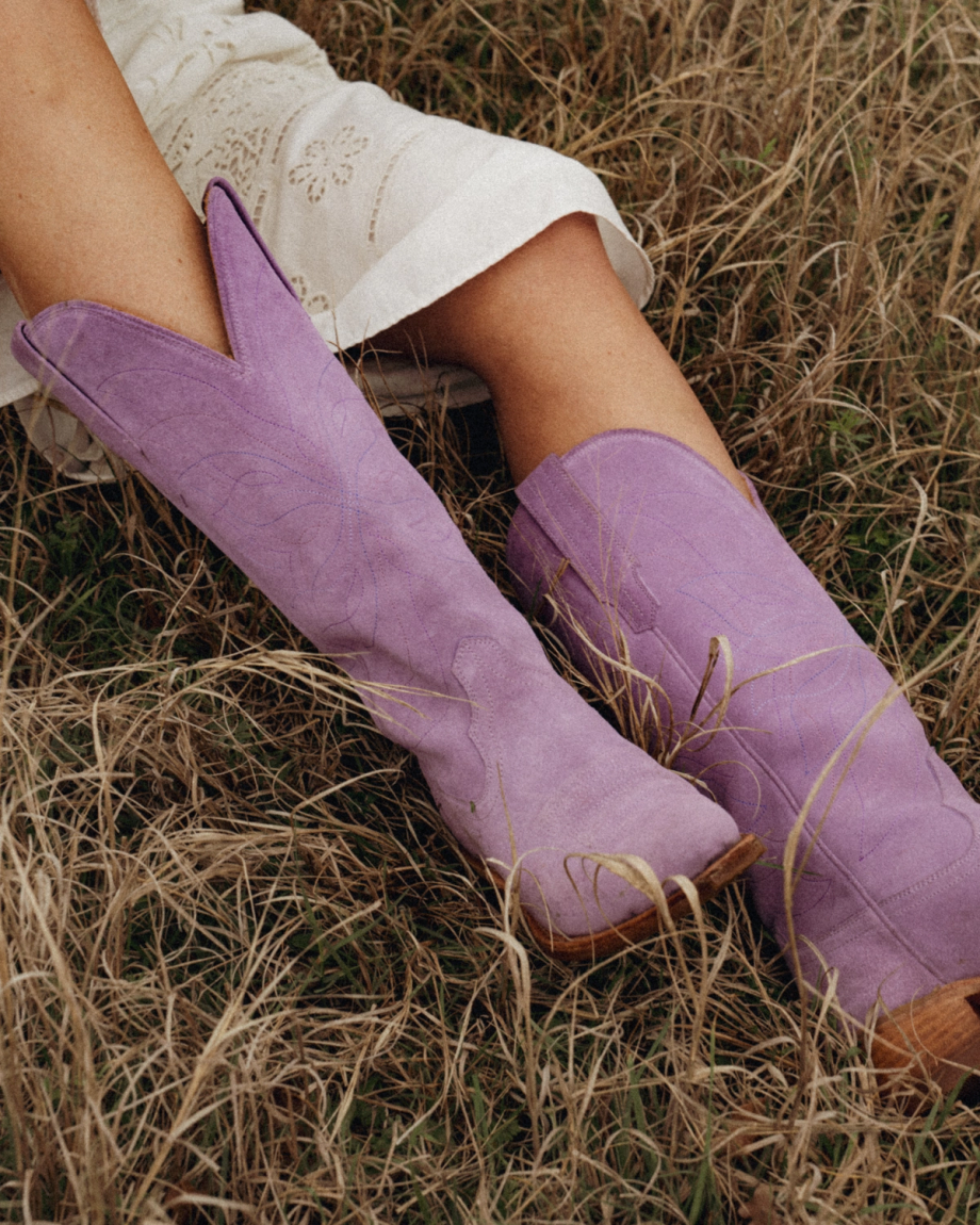 Person wearing a white embroidered skirt and The Annie Suede lavender cowboy boots sitting in dry grass.