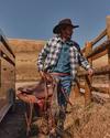 A man in a plaid shirt and cowboy hat carries a saddle near a wooden fence and trailer in a dry, grassy rural area.