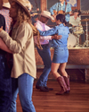 A couple in cowboy hats dances closely in a lively western-style bar with other dancers and a band in the background.