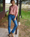 Woman stands on a wooden porch wearing a pink long-sleeve shirt, blue jeans, and tan boots. Grass, trees, and part of a building are visible in the background.