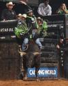 A bull rider wearing protective gear rides a bucking bull in an indoor rodeo arena, with spectators and event banners visible in the background.