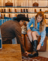 A man helps a woman try on a pair of cowboy boots in a store. The woman is seated on a leather seat in front of wooden shelves displaying various boots and bags.