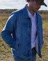 A person wearing a denim jacket, blue jeans, white shirt, and camouflage baseball cap stands in an open field. The background features a clear sky and a distant tree line.