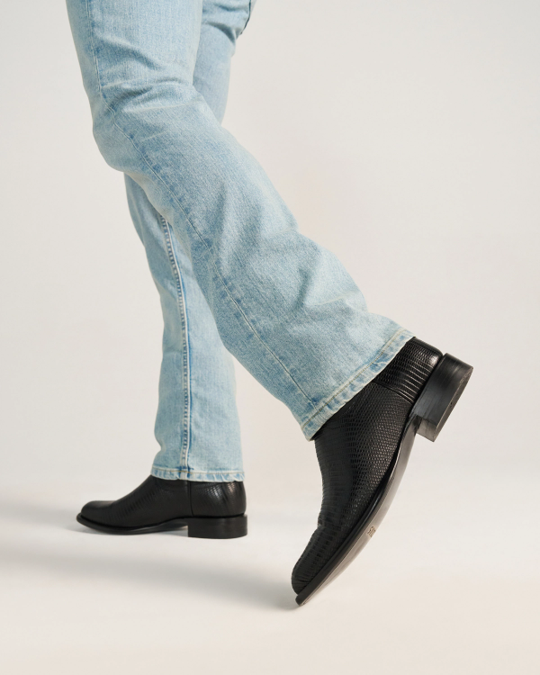 The Earl in Obsidian Lizard. Person wearing light blue jeans and The Earl Lizard black textured dress shoes, standing with one foot slightly forward on a plain light background.