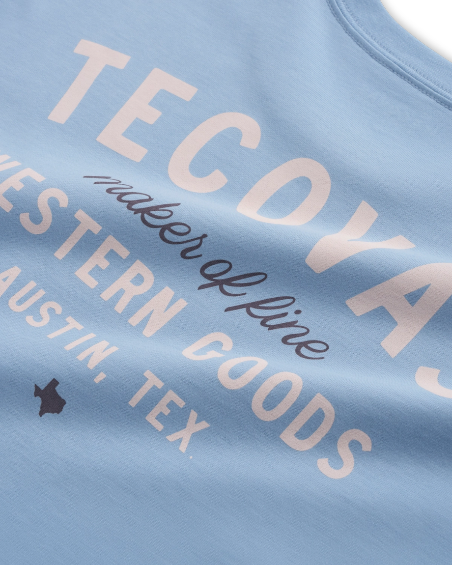 Close-up of a light blue shirt with white text reading "TECDYAS maker of fine WESTERN GOODS AUSTIN, TEX," plus a small Texas state outline icon.
