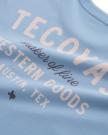 Close-up of a light blue shirt with white text reading "TECDYAS maker of fine WESTERN GOODS AUSTIN, TEX," plus a small Texas state outline icon.