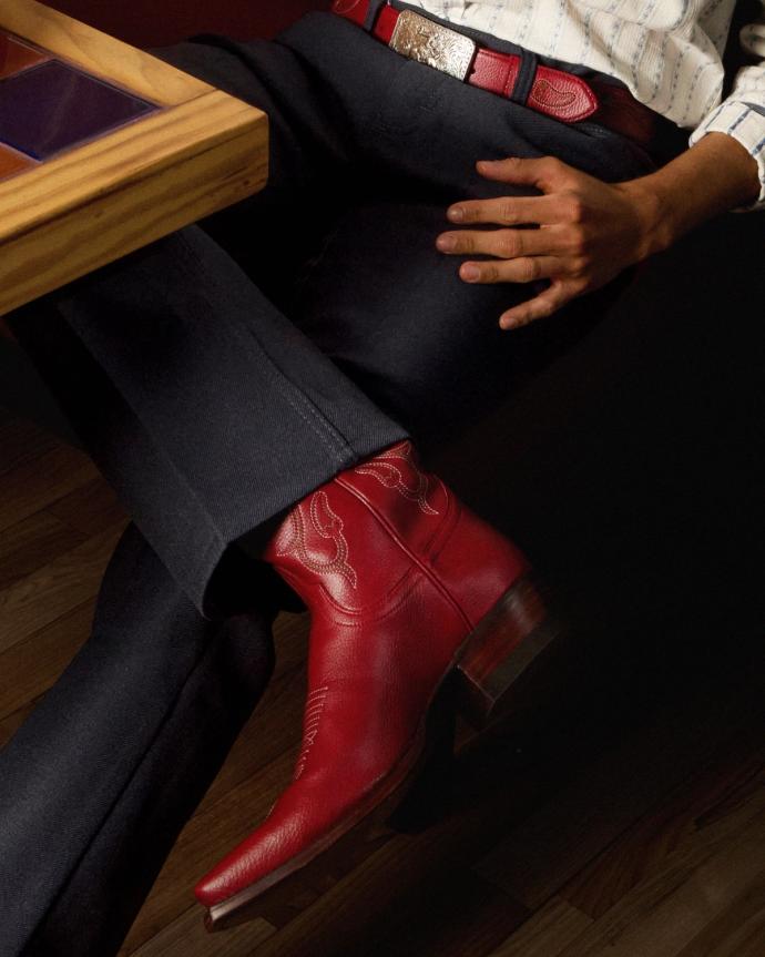 A person wearing dark pants, a red belt with a silver buckle, and red cowboy boots sits with legs crossed beside a wooden table.