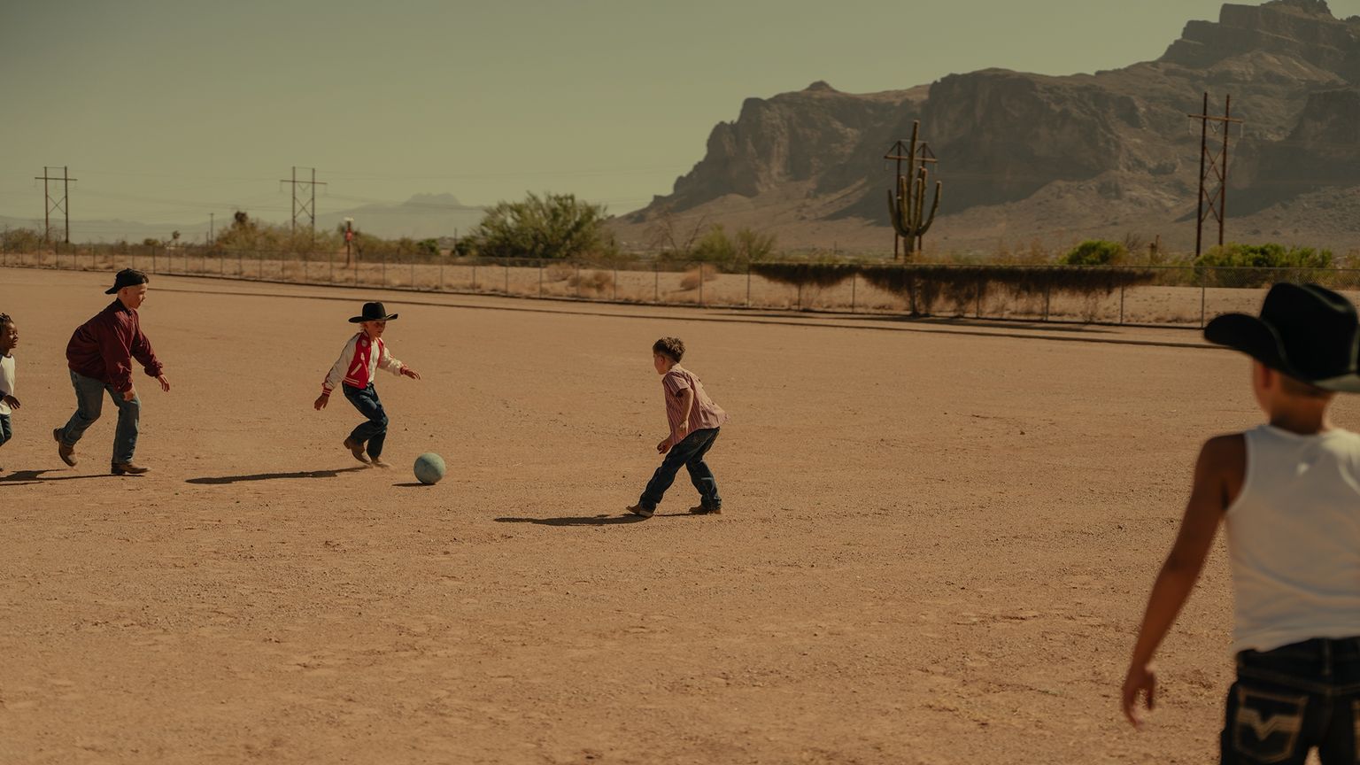 Five children, some wearing cowboy hats, play soccer on a dusty field with mountains and desert vegetation in the background.