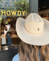 A woman wearing a beige cowboy hat with "rh" initials stands in front of a store window with the word "howdy" displayed.