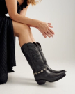 A person in a black dress sits with legs crossed, wearing black cowboy boots with decorative stitching and silver accents.